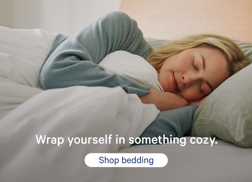 Woman sleeping under a blanket with promotional text that says, "Wrap yourself in something cozy. Shop bedding."