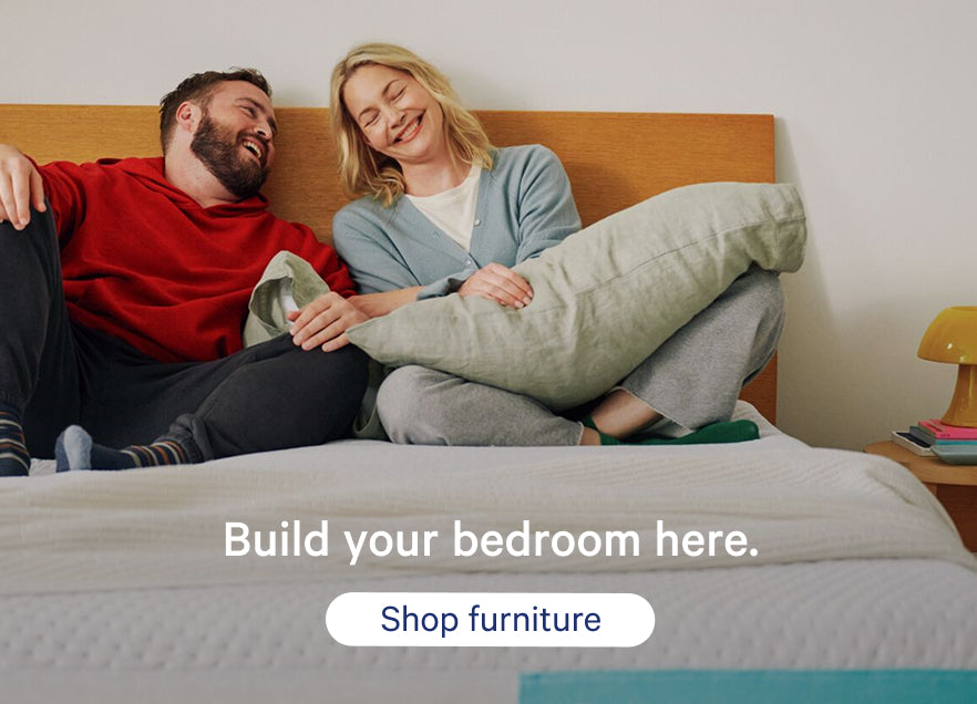 A couple sits on their mattress against a wooden headboard. Promotional text on the image says, "Build your bedroom here. Shop furniture."