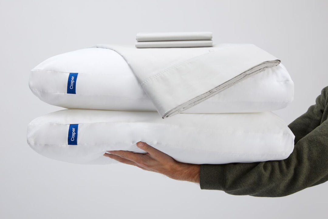 A person holds two pillows topped with folded pillowcases and a sheet set.