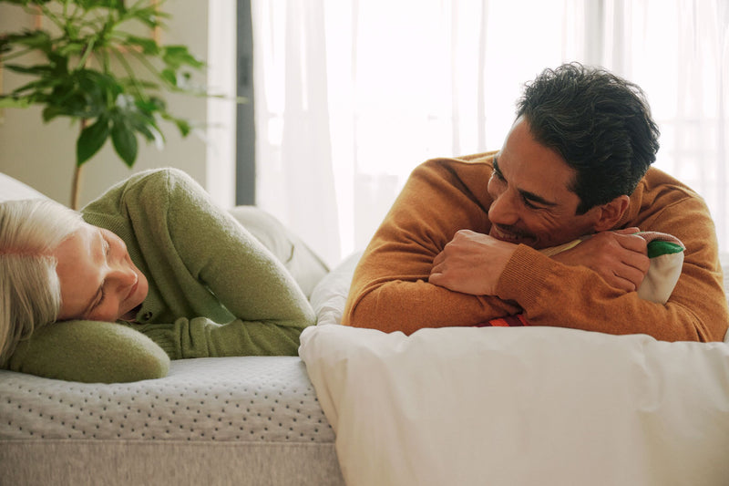 A couple lies down, smiling at each other, on their mattress.
