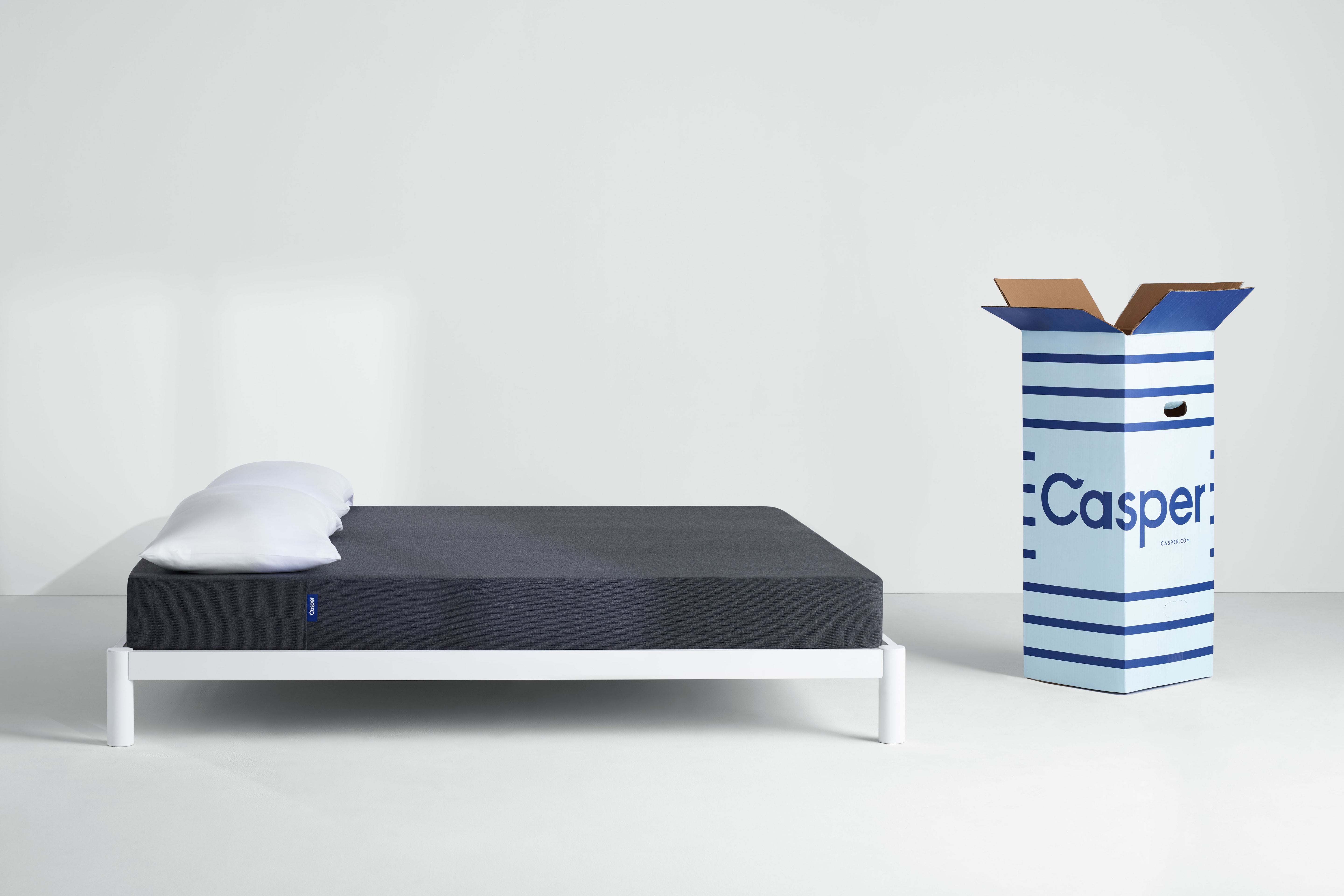 Casper's Essential 2025 mattress, side view, with its Casper shipping box standing opened next to the bed.