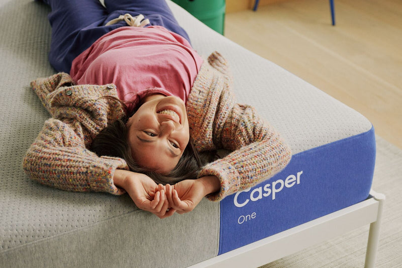 A person on their back smiles while lying near the edge of The One mattress