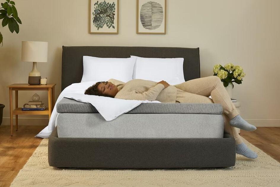Front view of a mattress in a bedroom with a mattress topper on it. A woman lies comfortably on her back on the mattress topper.