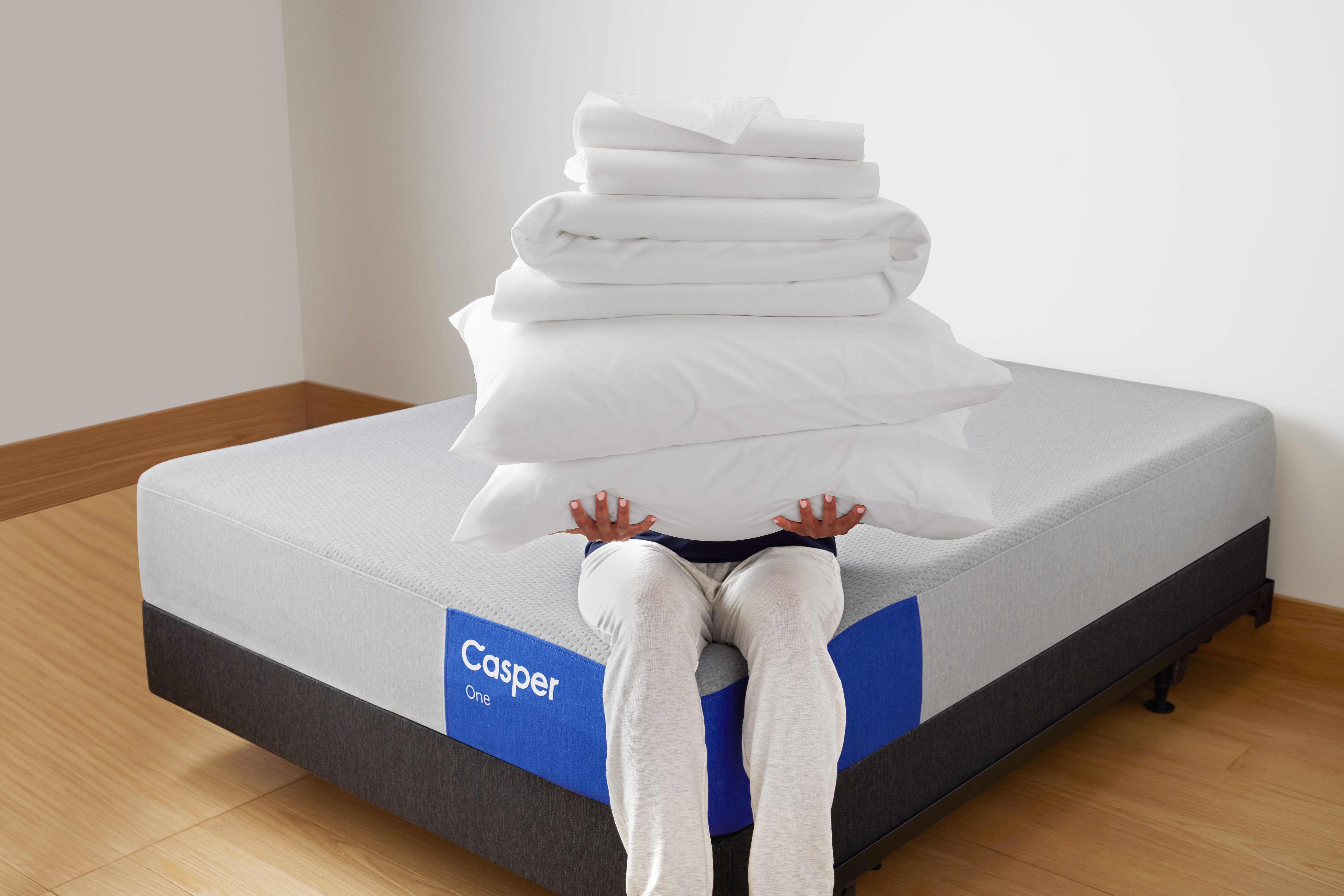 A person sits on the corner of a Casper One mattress holding a stack of products including two pillows, a folded mattress protector, and a folded sheet set.
