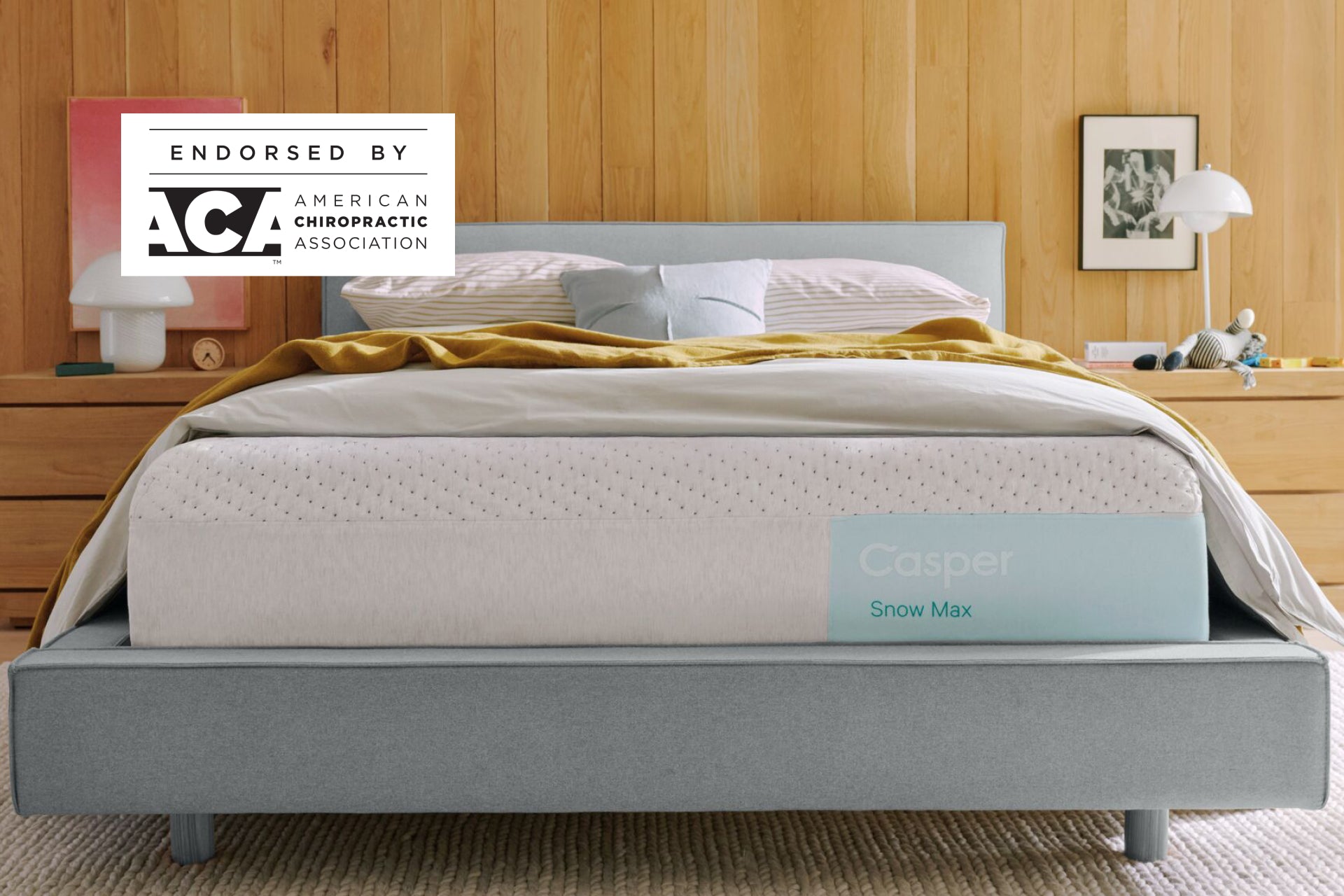 Casper's Snow Max mattress on a bed frame with pillows and a blanket folded on top. A badge on the image indicates that this mattress is endorsed by the American Chiropractic Association.