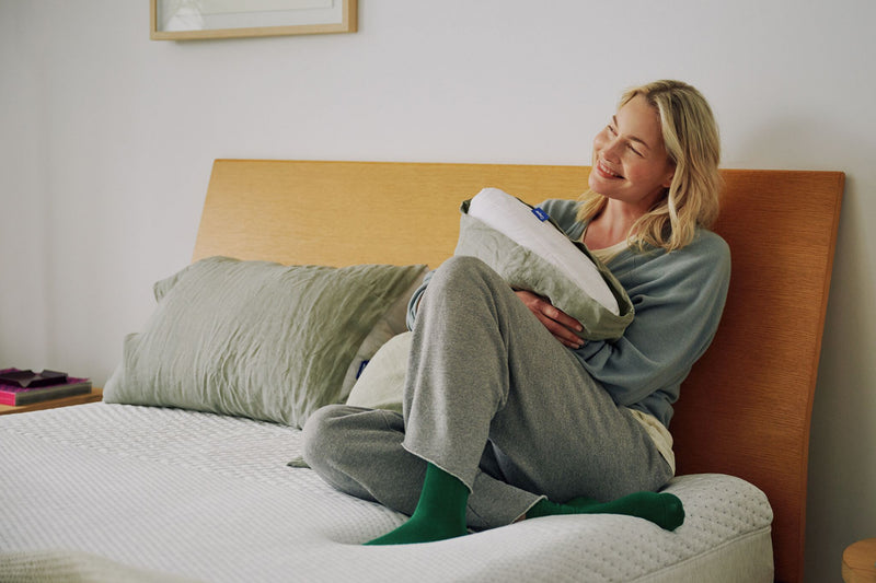 A woman sits on her mattress against a wooden headboard.