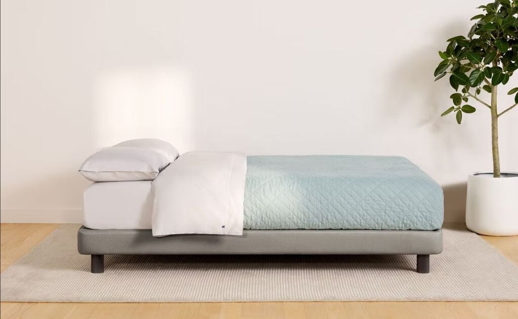 Side image of a fully made bed against an empty wall. Casper's Upholstered Bed Frame has a mattress on it and a large, thin rug underneath it.