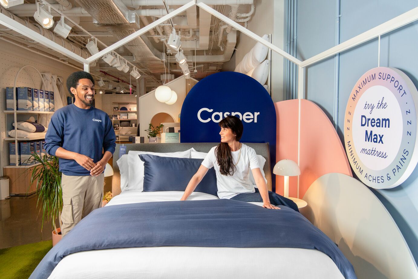 One person standing next to a made bed with another sitting on it, both smiling.